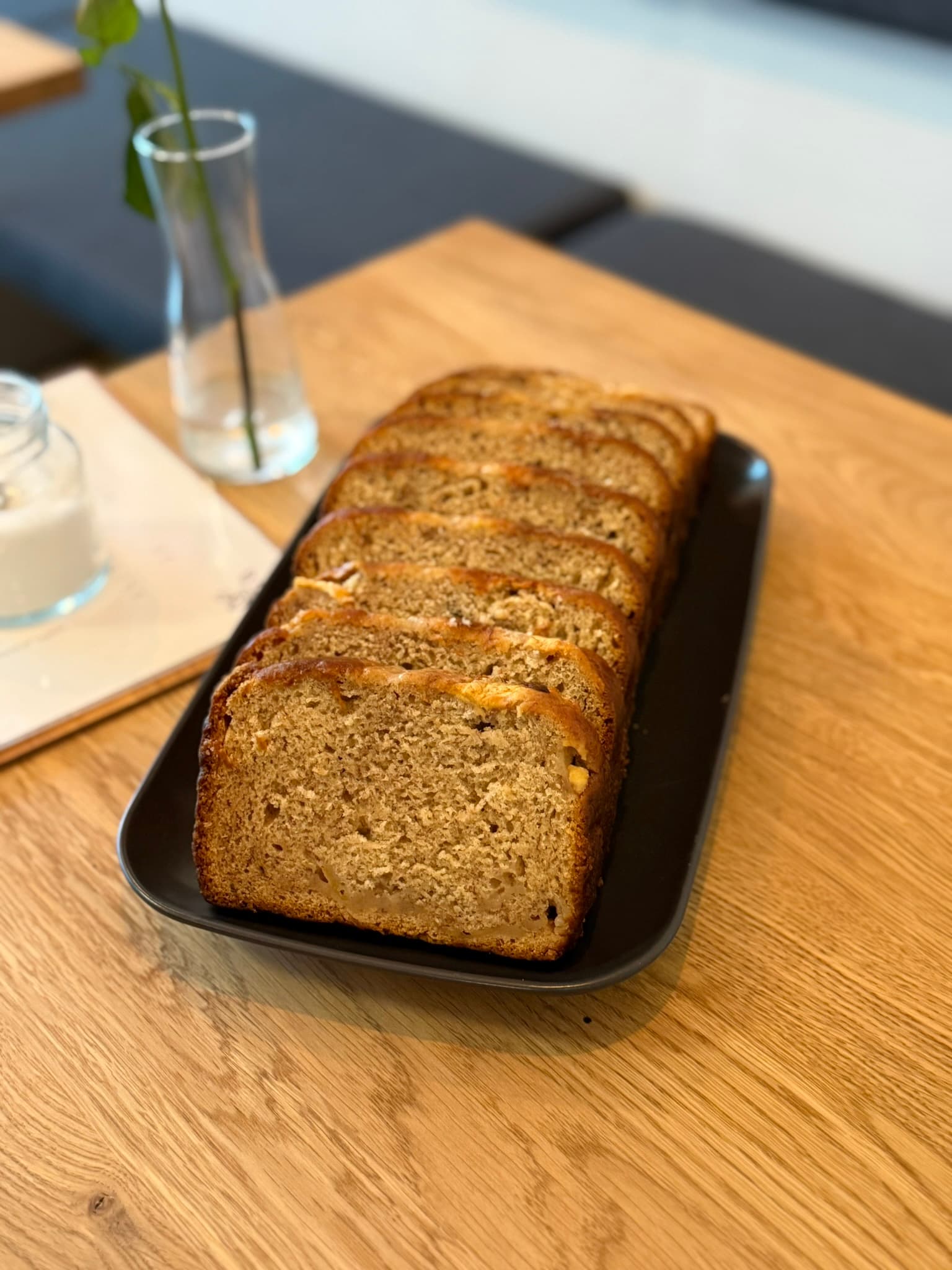 Klassisches Bananenbrot von de Bananas in Scheiben auf schwarzer Schale, Tageslicht, Blumenvase im Hintergrund