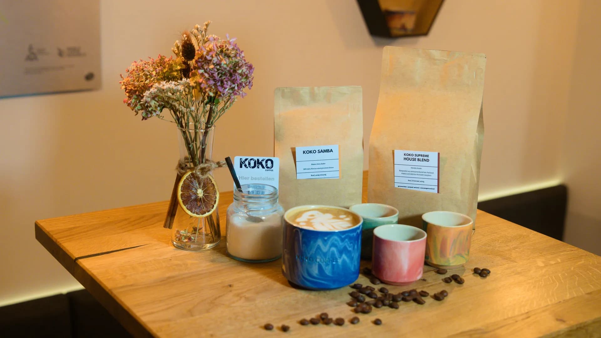 KOKO COFFEE LAB Produktarrangement – Bohnentüten Samba und Supreme House Blend mit Keramiktassen, Latte Art und Blumenstrauß auf Holztisch