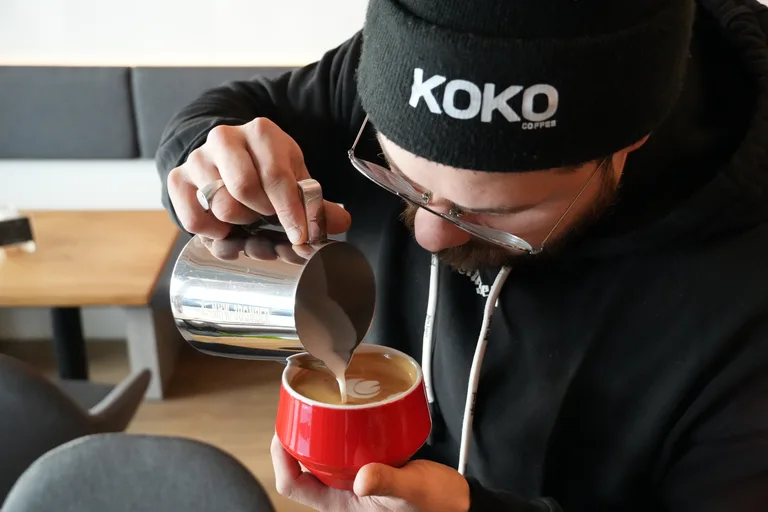 Barista mit KOKO-Coffee-Beanie gießt Latte Art in eine rote handgemachte Keramiktasse im KOKO Coffee Lab Würzburg