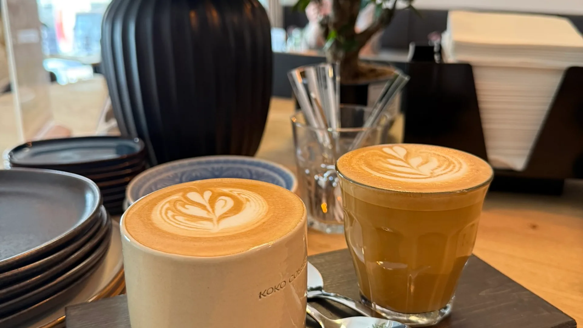 Cappuccino in Keramiktasse und Cortado im Glas nebeneinander auf Serviertablett, Café-Interior mit Flavor-Wheel an der Wand