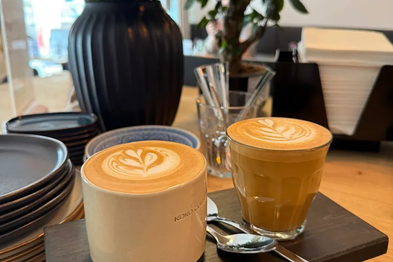 Cappuccino in Keramiktasse und Cortado im Glas nebeneinander auf Serviertablett, Café-Interior mit Flavor-Wheel an der Wand