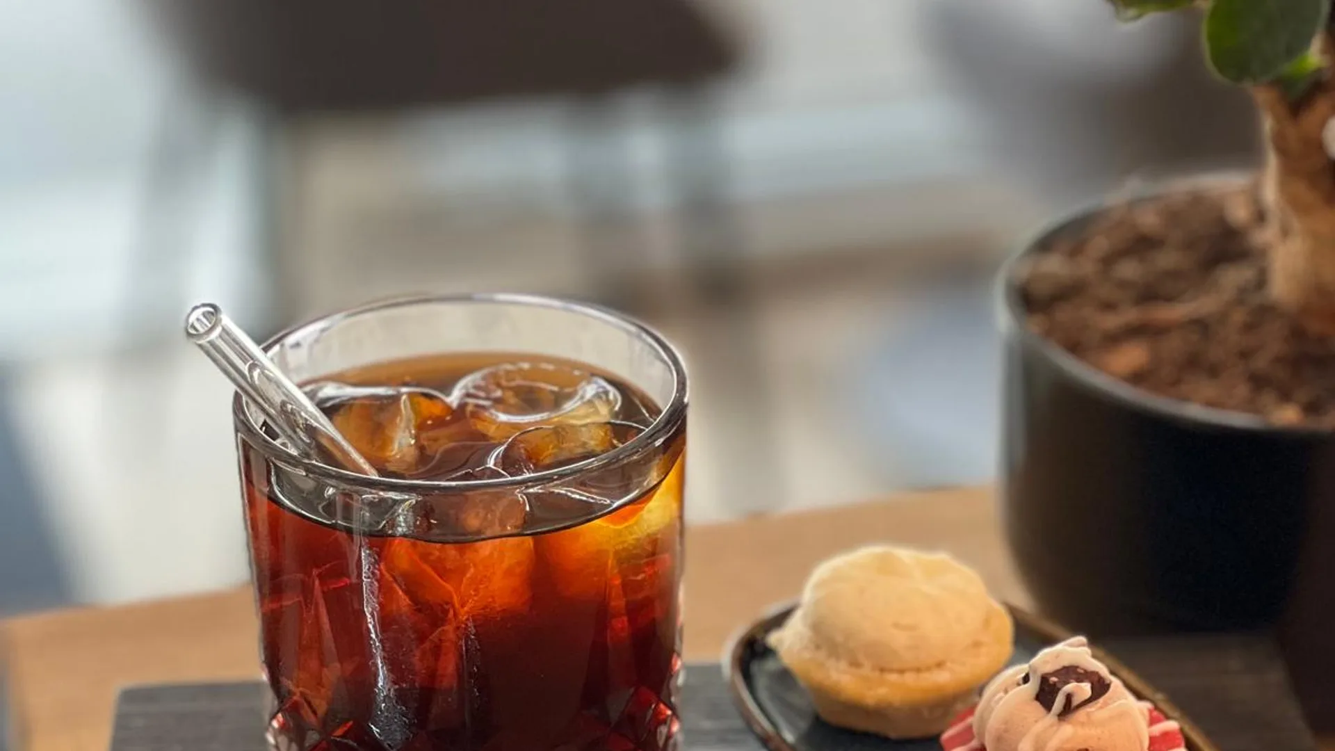 Cold Brew im Whiskyglas mit Eiswürfeln und Glas-Rührstab auf dunklem Holzbrett, daneben Teller mit kleinen Bakes, Bonsai im Hintergrund