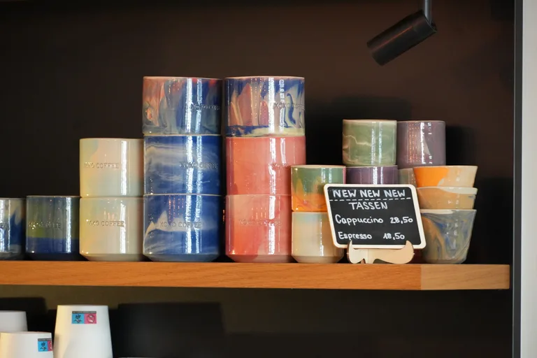 Bunte handgemachte Keramiktassen im Holzregal des KOKO Coffee Lab vor dunkler Wand – Cappuccino- und Espressotassen zum Kauf