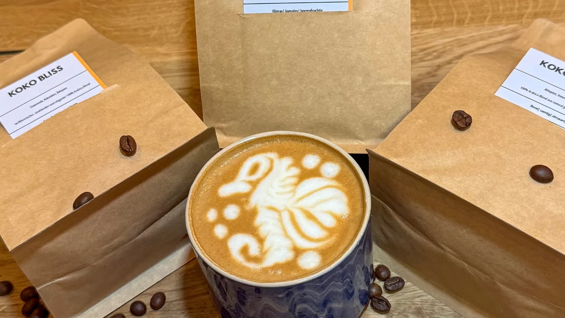Drei KOKO Bohnentüten – Harmony und zwei Bliss – arrangiert um Latte in blauer Keramiktasse, Kaffeebohnen verstreut