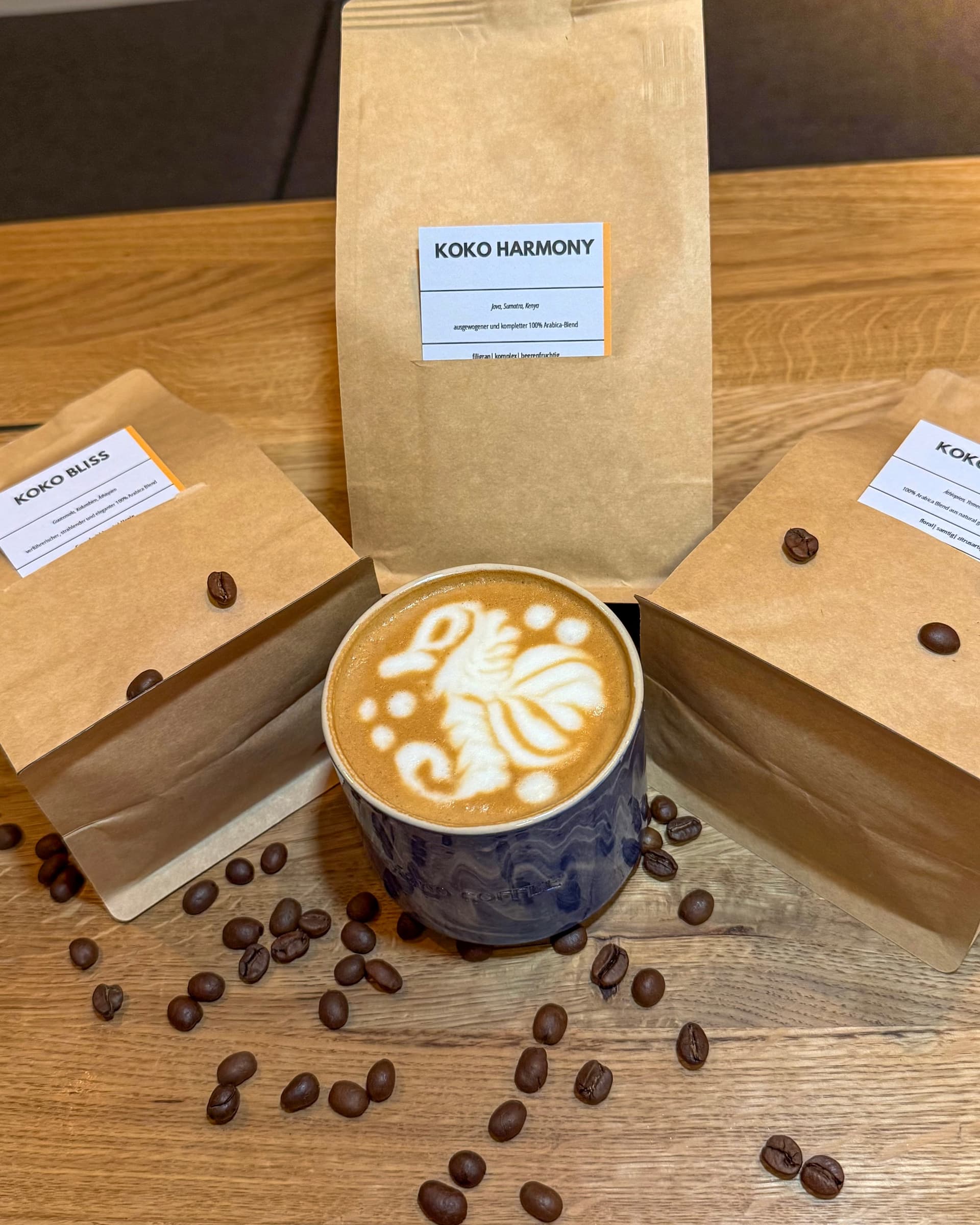 Three KOKO bean bags – Harmony and two Bliss – arranged around latte in blue ceramic cup, coffee beans scattered