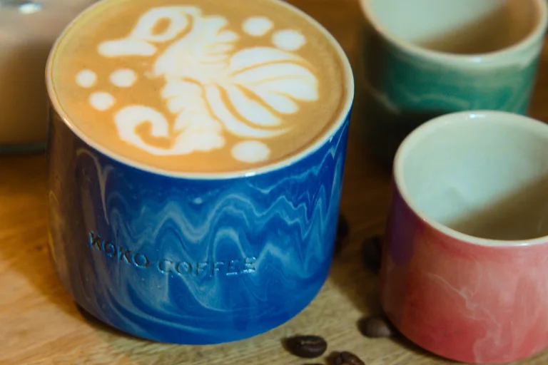 Latte mit floraler Latte Art in blauer Wellenglasur-Keramiktasse von KOKO Coffee, umgeben von frisch gerösteten Kaffeebohnen