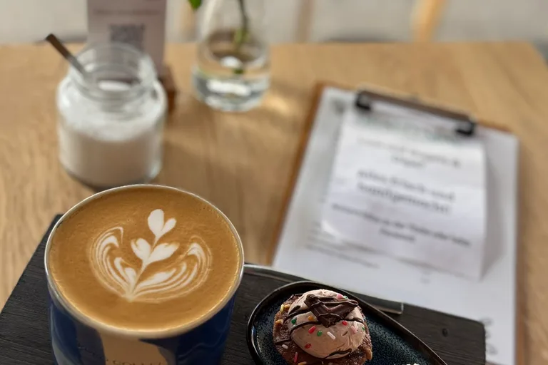 Latte in blauer Keramiktasse mit Latte Art und kleinem Dessert auf Holzbrett, rote Rose in Vase