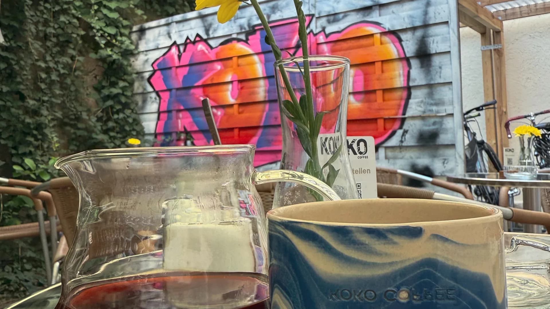 Outdoor-Tisch im KOKO Coffee Lab mit Pour-Over-Karaffe und blauer Keramiktasse, KOKO-Graffiti an der Wand