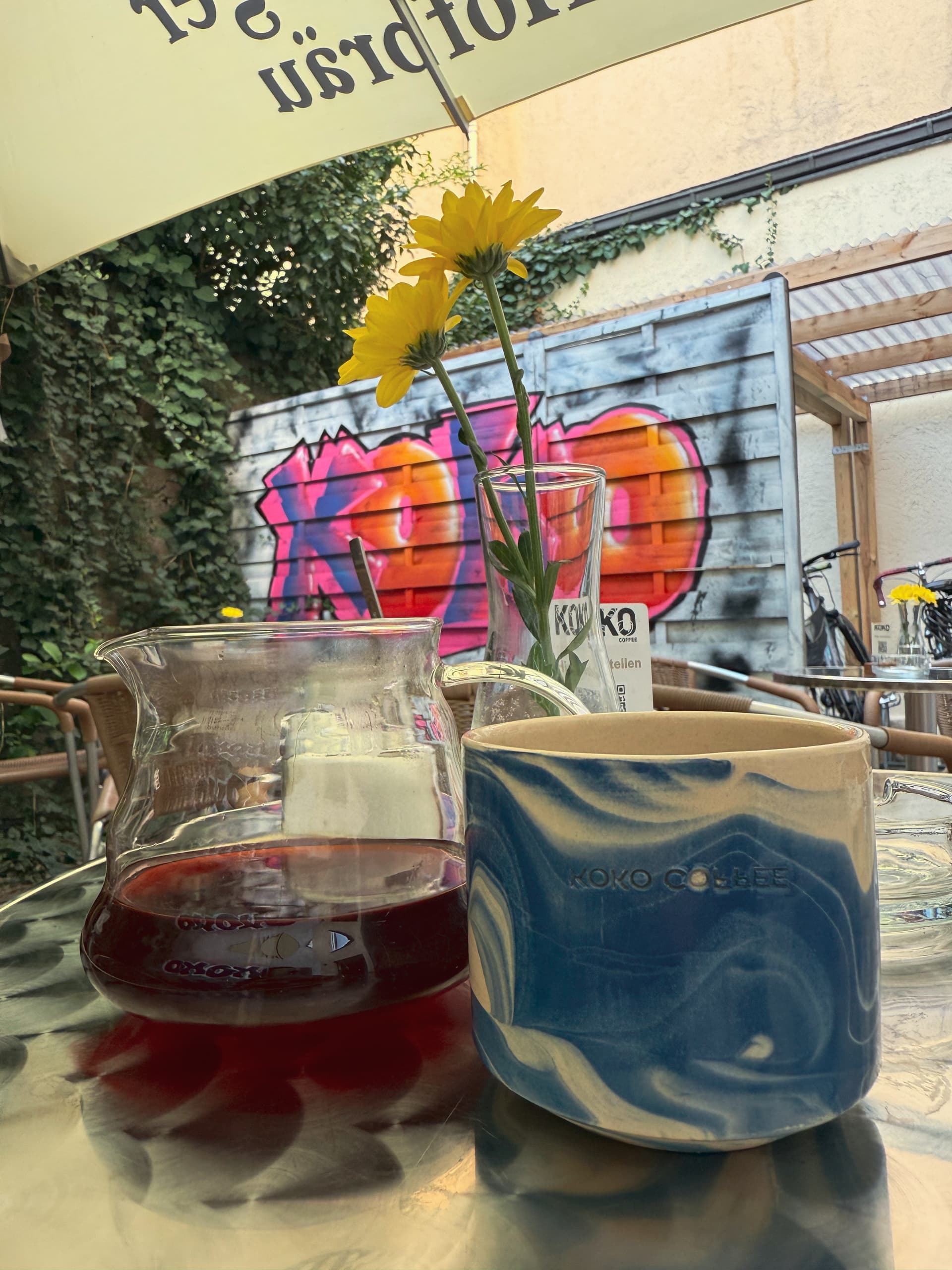 Outdoor table at KOKO Coffee Lab with pour-over carafe and blue ceramic cup, KOKO graffiti on wall