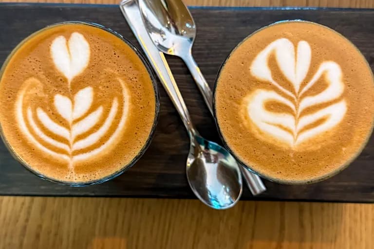 Zwei Cortados mit Latte Art in Glas-Tassen auf dunklem Holzbrett, von oben fotografiert