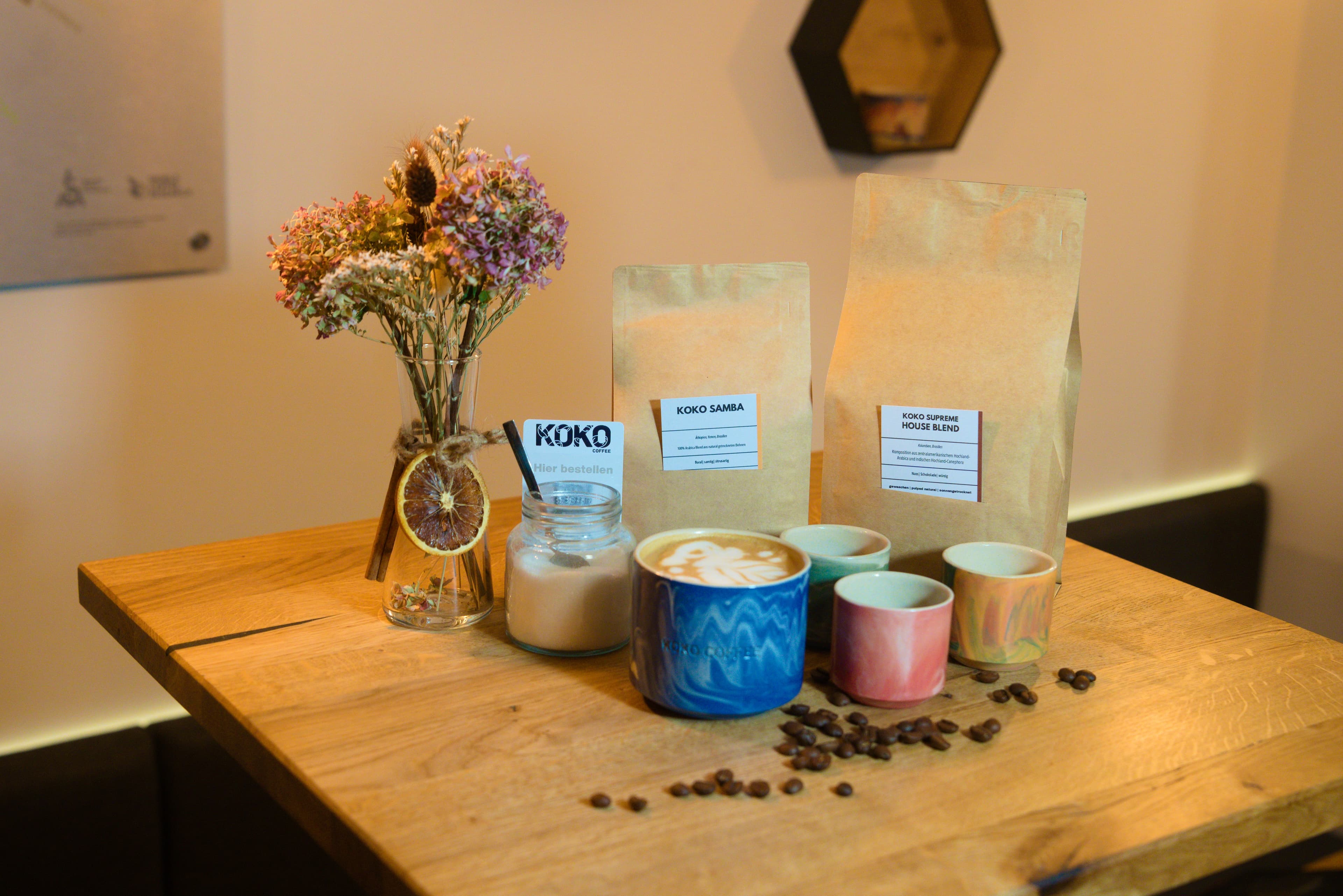KOKO COFFEE LAB Produktarrangement – Bohnentüten Samba und Supreme House Blend mit Keramiktassen, Latte Art und Blumenstrauß auf Holztisch