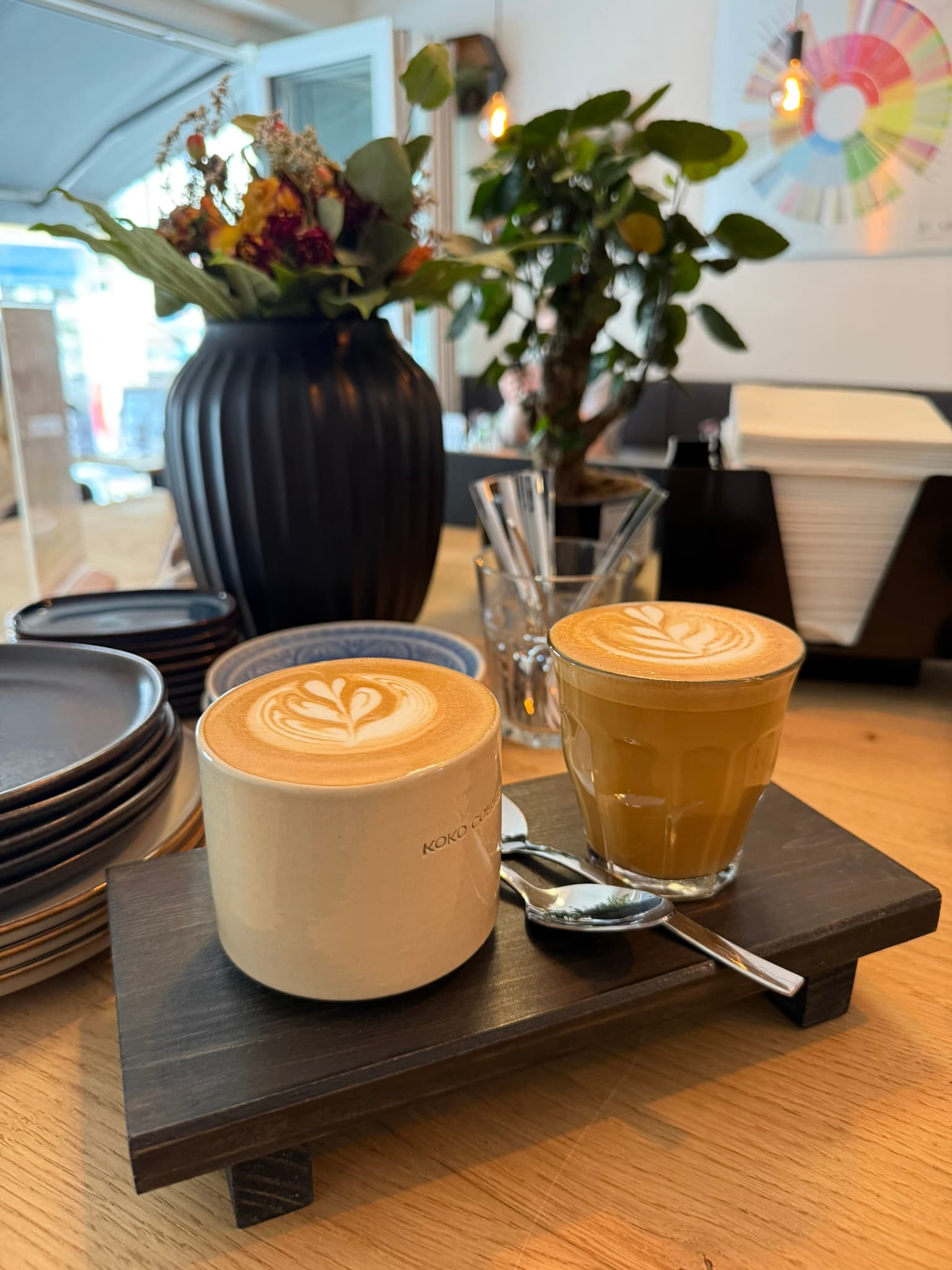 Cappuccino in Keramiktasse und Cortado im Glas nebeneinander auf Serviertablett, Café-Interior mit Flavor-Wheel an der Wand