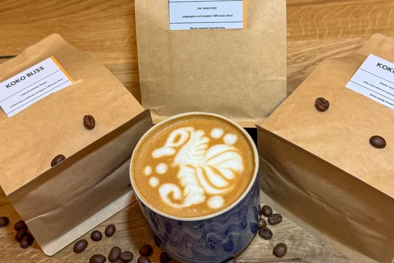 Drei KOKO Bohnentüten – Harmony und zwei Bliss – arrangiert um Latte in blauer Keramiktasse, Kaffeebohnen verstreut