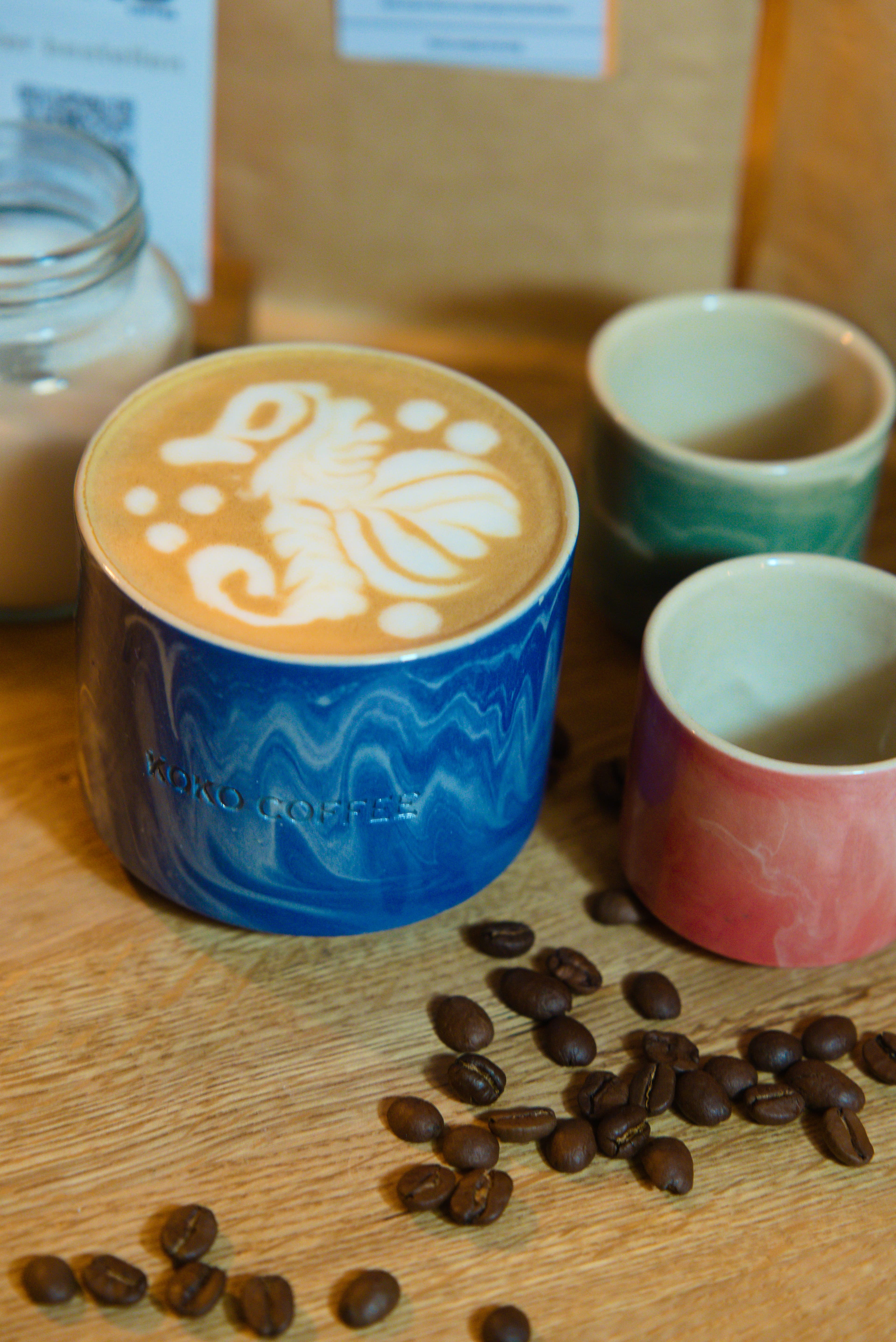 Latte mit floraler Latte Art in blauer Wellenglasur-Keramiktasse von KOKO Coffee, umgeben von frisch gerösteten Kaffeebohnen