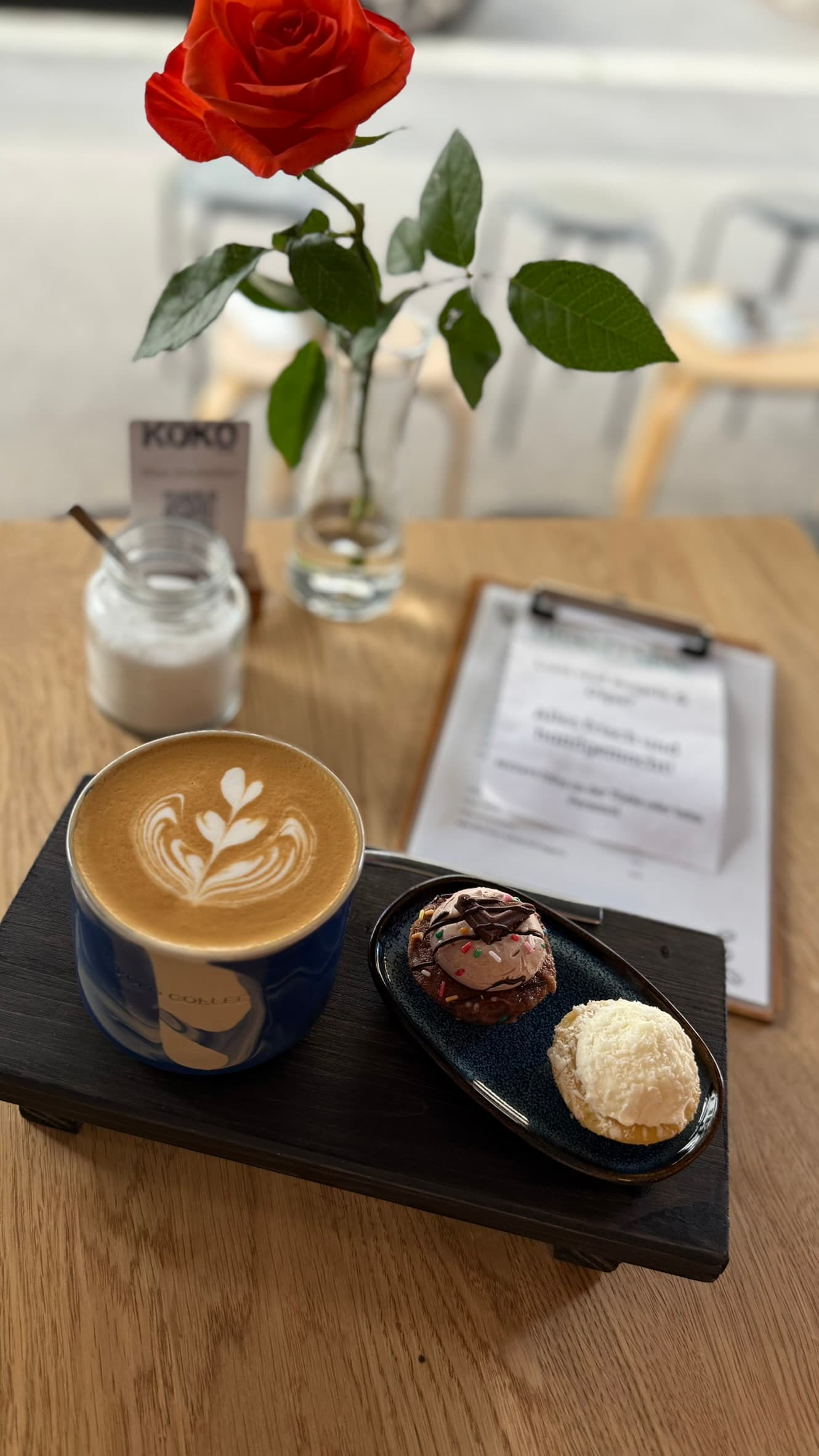 Latte in blauer Keramiktasse mit Latte Art und kleinem Dessert auf Holzbrett, rote Rose in Vase